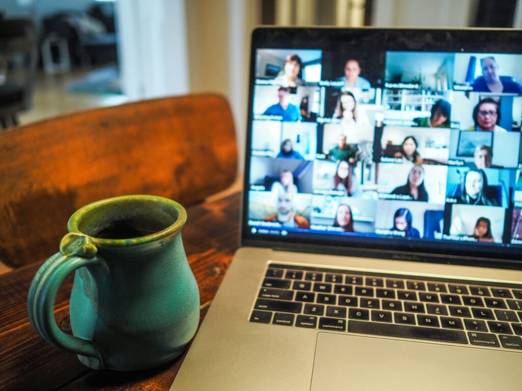 Close-up of a laptop on a wooden table displaying a video conferencing grid with multiple participants, each in their own video window. A green ceramic mug sits to the left of the laptop. The scene depicts a remote learning or virtual meeting environment, with participants engaged in an online class or discussion