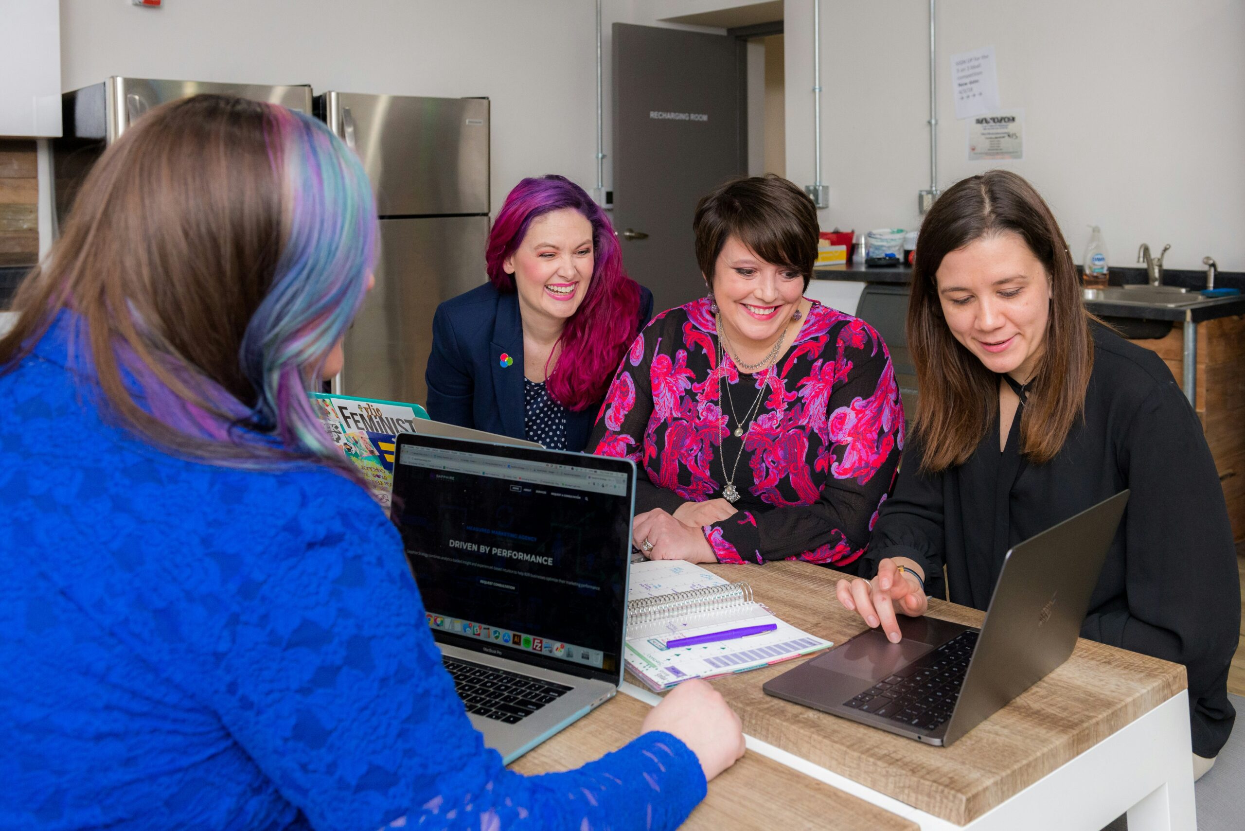 Four individuals seated around a wooden table, in discussion with two open laptops, notebooks, and pens. One person points at a laptop screen while others listen. The background includes a kitchenette with stainless steel appliances