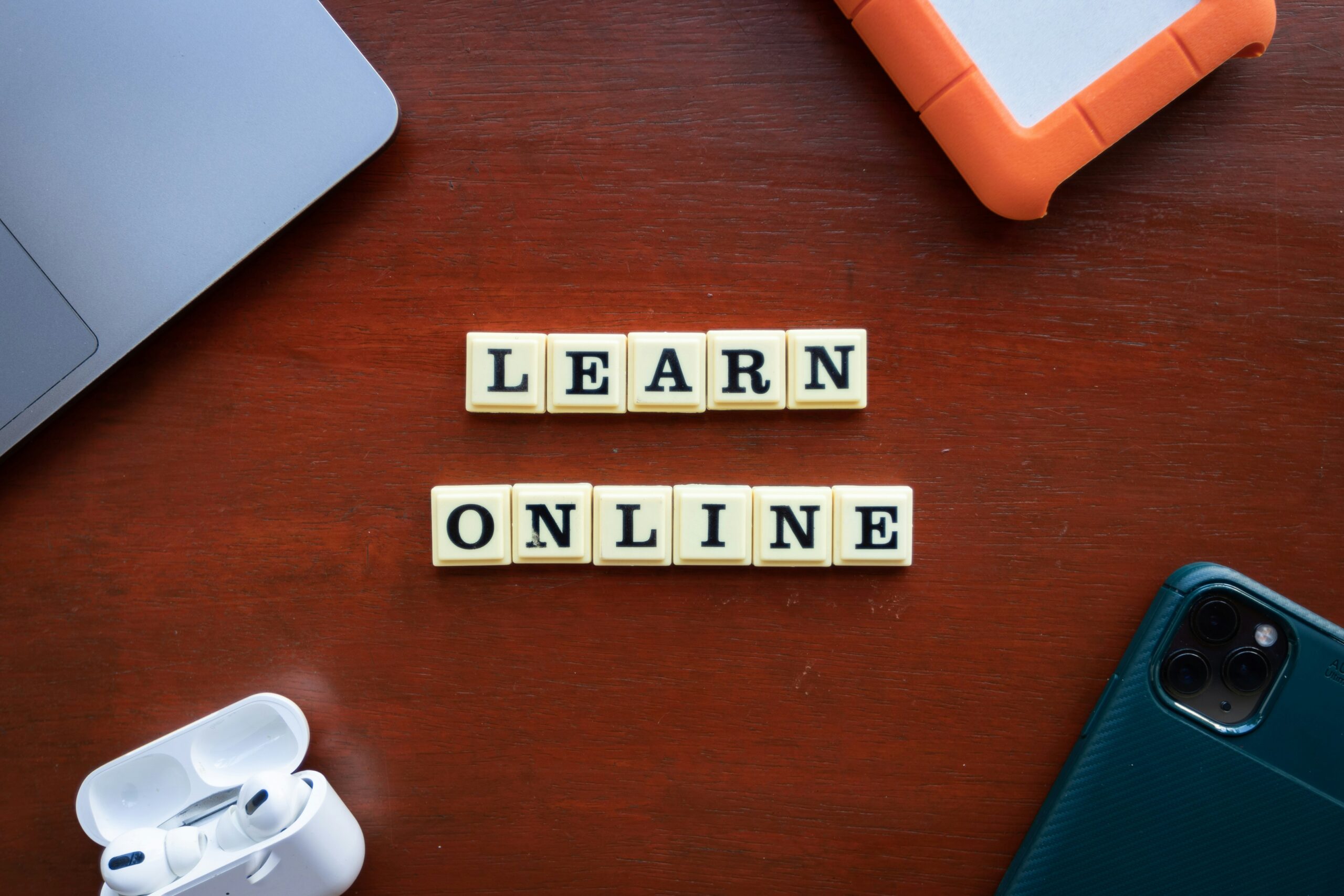wooden surface with Scrabble-style tiles spelling 'LEARN ONLINE' in two rows. Surrounding the tiles are a partially visible laptop, a pair of wireless earbuds with their case, an external hard drive with an orange bumper, and a phone in a green case