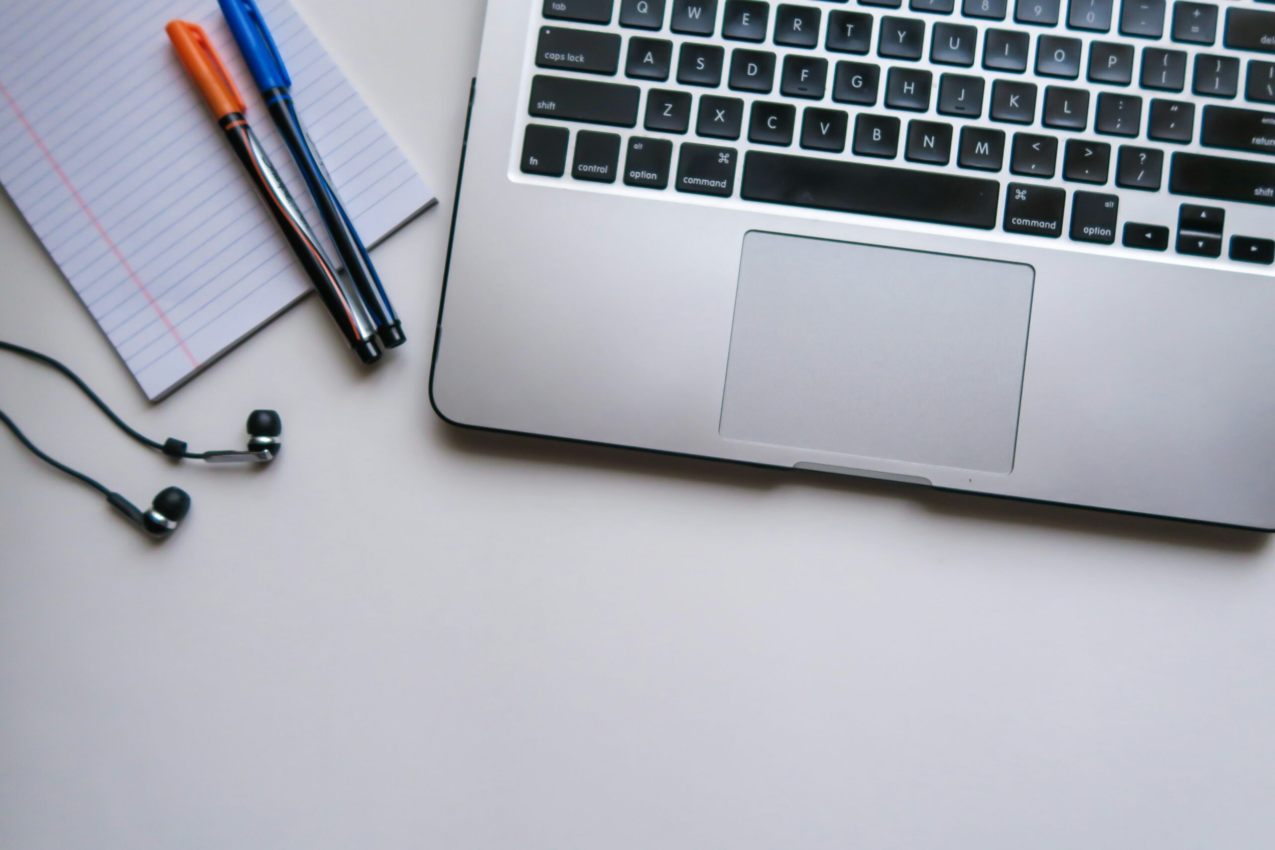 workspace setup featuring the corner of a laptop keyboard, a lined notepad with a blue and an orange pen resting on top, and a pair of black wired earphones placed below the notepad. All items are arranged on a clean white surface