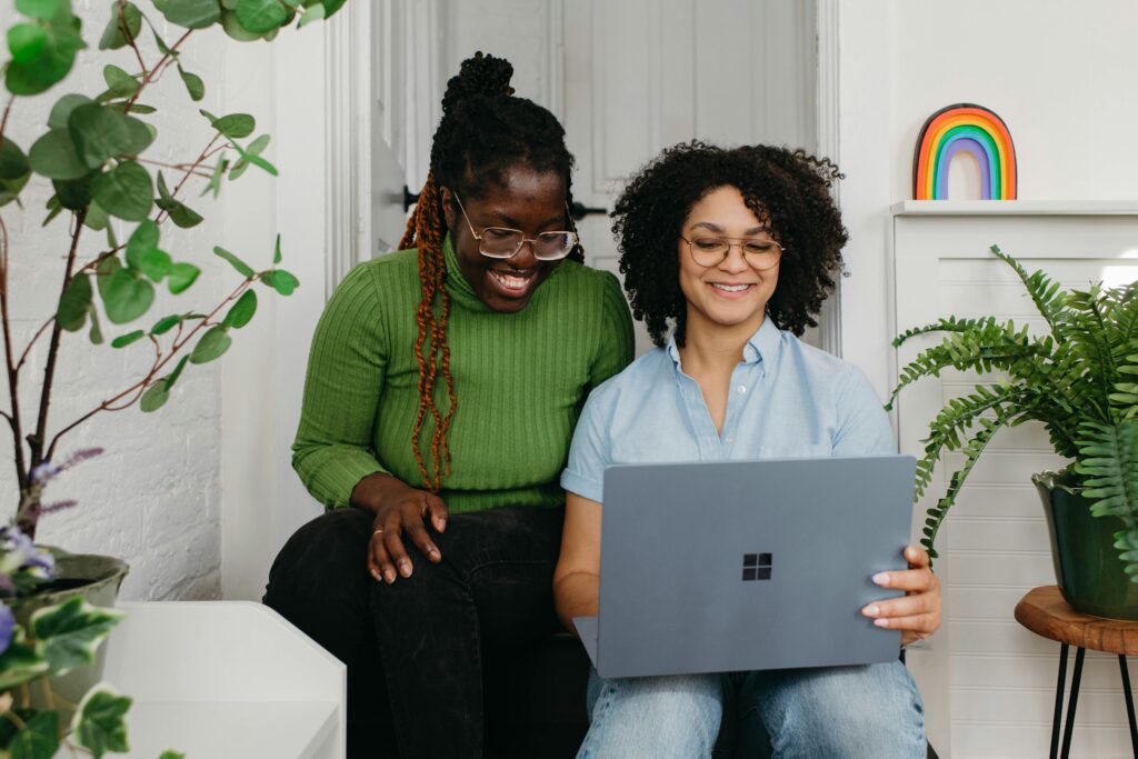 Two individuals sitting indoors, focused on a laptop. One wears a green sweater and black pants, the other a light blue button-up shirt and jeans. Both wear glasses and appear engaged in a shared task. The setting includes green plants, a white wall, and a shelf with a rainbow decoration